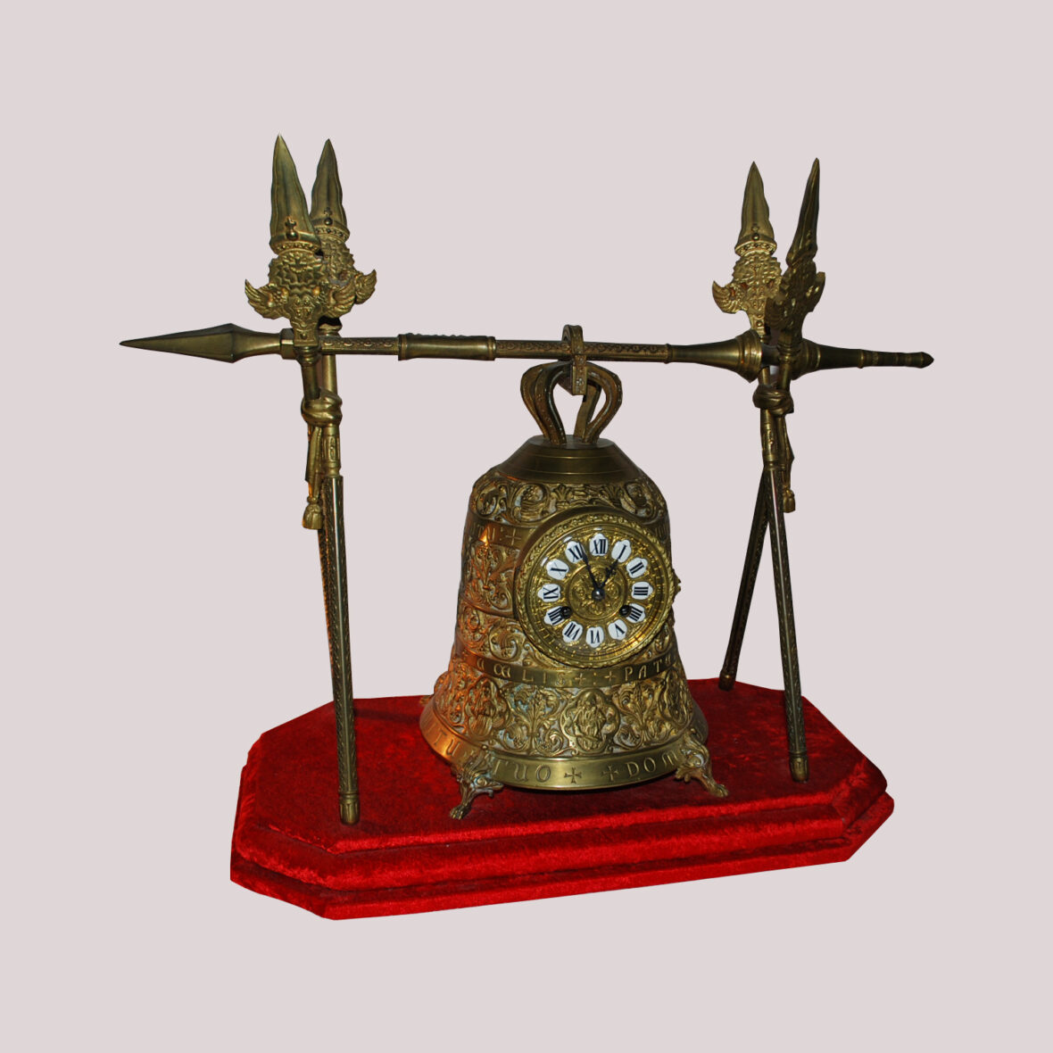 Pendule en bronze doré représentant une cloche tenue par des lances, le cadran à médaillon émaillé blanc et chiffres romains. Socle en velours rouge. De style gothique.
